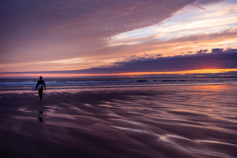 a surfer in a wetsuit walking across a beach at sunset towards the sea with the sun behind a cloud causing a purple sky, beach sunset heavy clouds 4k