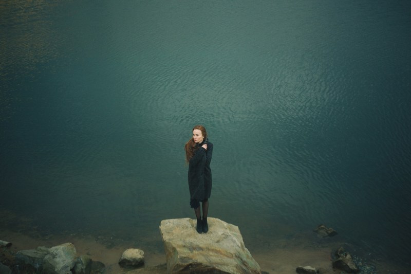 a redheaded woman bundled in a black sweater and black boots standing on a rock next to a lake, the girl by the lake 4k
