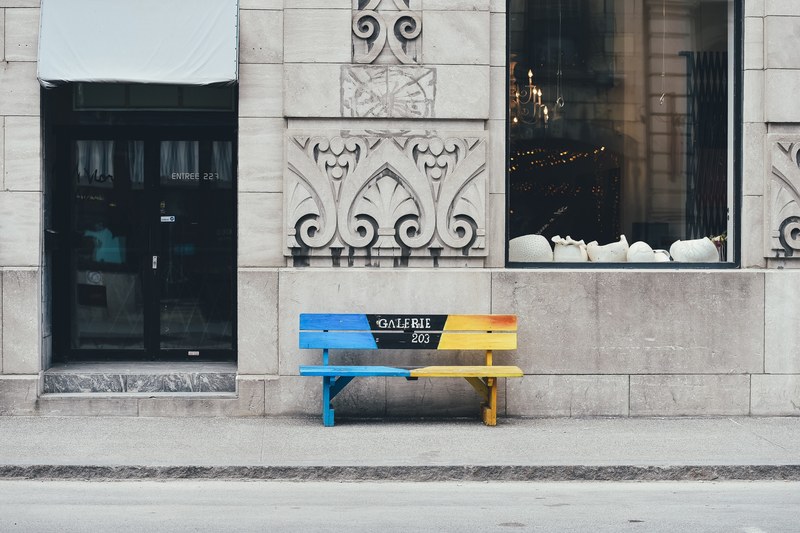 a blue and yellow short wooden bench located on a sidewalk outside a building in old montreal, bench on a sidewalk_ 4k