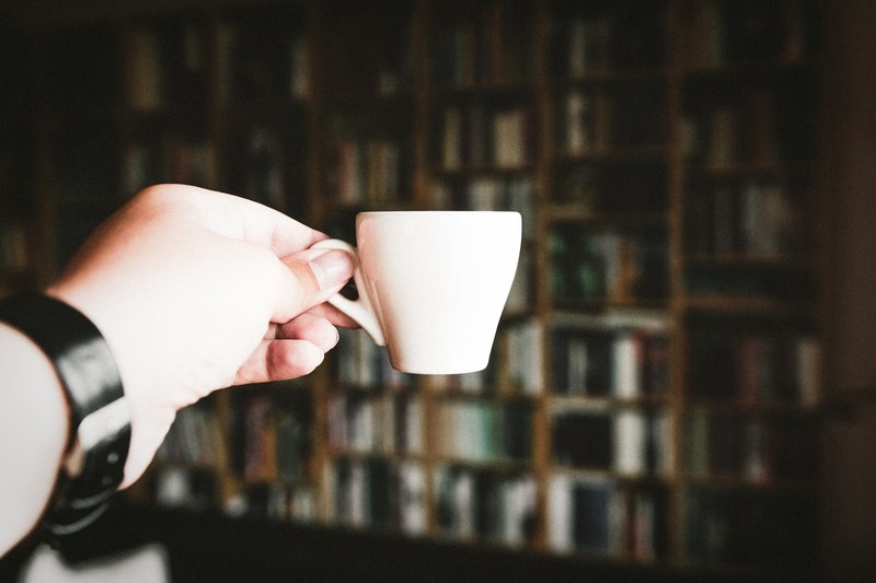 persons hand with a watch holds a small espresso cup in front of bookshelves and books, the goal_ 4k