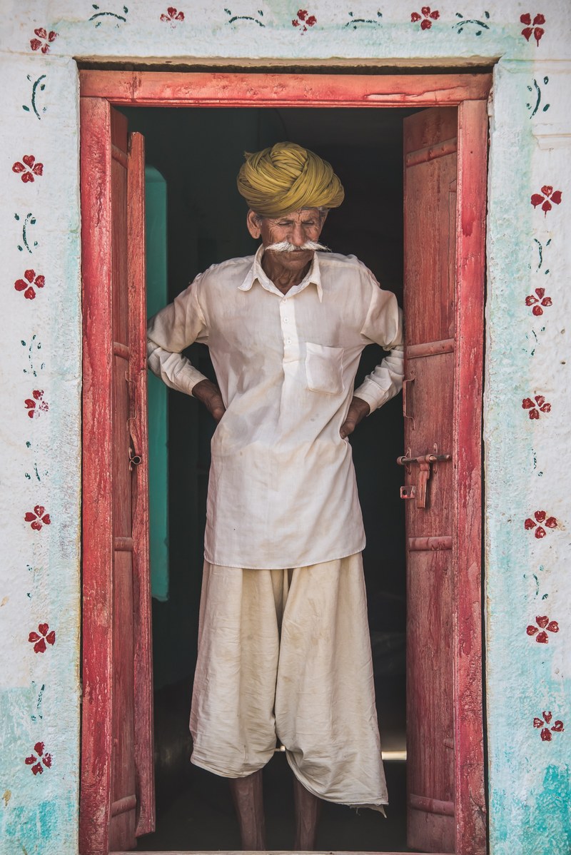 indian man with flowy clothes and a yellow turban standing in a pink doorway with flower on the walls, indian man standing in doorway 4k