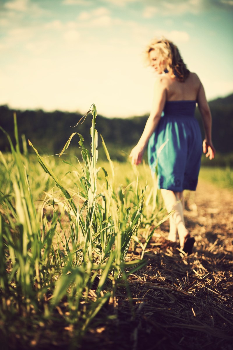 a blonde woman with a blue dress walking down a path of hay looking at the field of growing tallgrass, countryside walks 4k