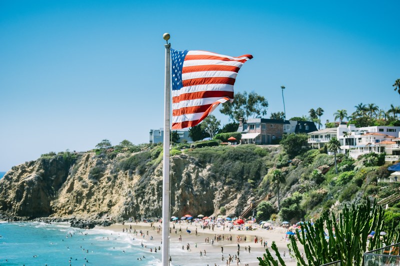 the american flag flutters in the wind over a crowded beach by a cliff, flag over a beach 4k