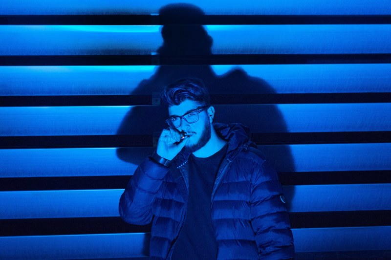man wearing glasses with a beard smoking a cigarette wearing a puffy jacket inside a garage lit with a green light, blue guy smoking 4k
