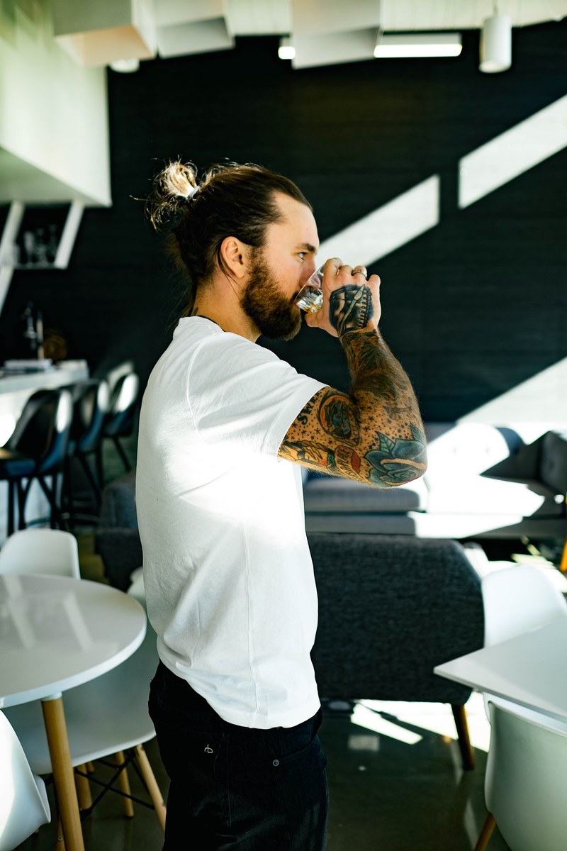 man with colorful arm tattoos taking a sip from a glass while standing in a restaurant, tattooed man drinking 4k