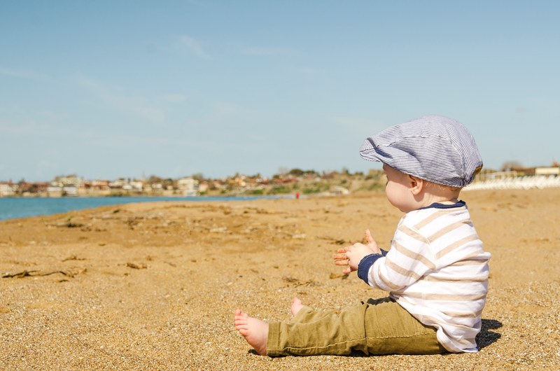 a baby boy wearing a blue stripped hat sits on the sand clapping as he looks towards the ocean, _a baby boy sitting on the sand 4k
