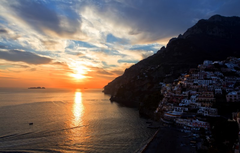 a cloudy orange sunset reflecting on the water by an oceanside town built on a hill, positanos sunset 4k
