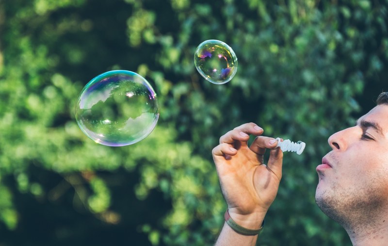 man holding and blowing into a bubble wand and blowing out two bubbles in front of a bush, _blowing bubbles 4k