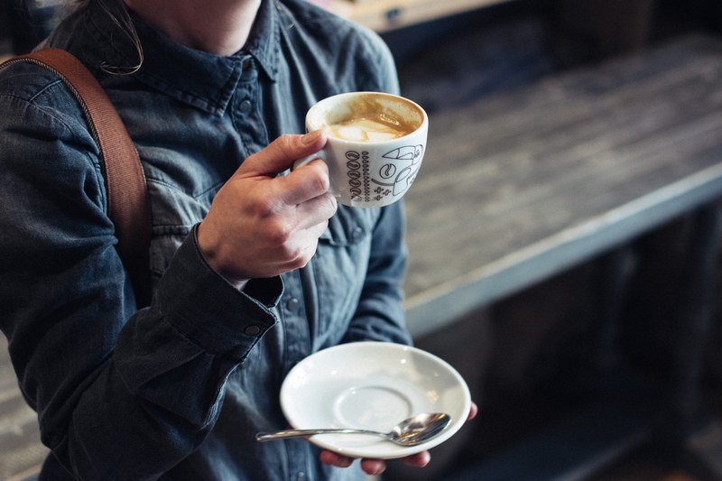 a woman in buttoned up denim shirt with a handbag lifting a mug of cappuccino in one hand and holding a saucer and teaspoon in another, _woman holding her cup of coffee with milk 4k