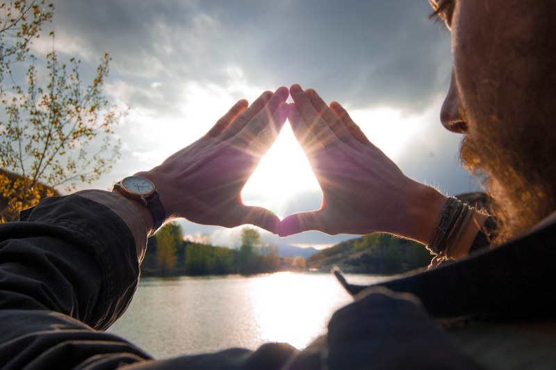 a bearded man beside a lake holds his hands in a triangle the sun shining through, sun shining through hands 4k