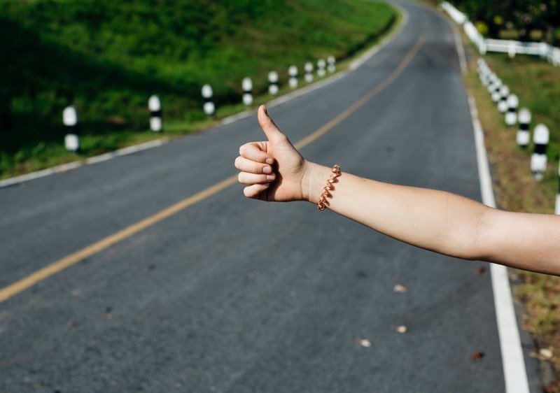 persons arm with bracelet displaying hitch hiking sign near asphalt road, hitch hiking sign near road 4k