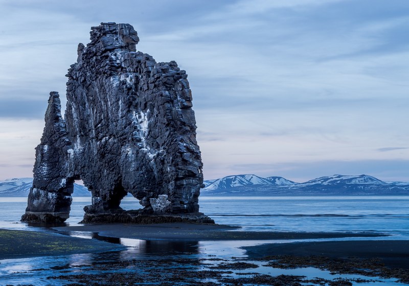 tall rock formation above the ocean with snow covered mountains in the background at hvtserkur, rock formation above the ocean 4k