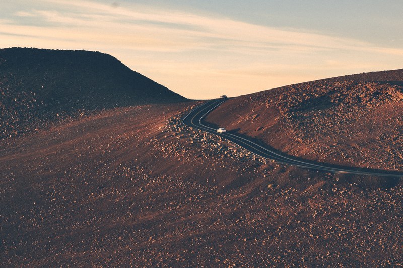 two cars driving up an asphalt road on a slope covered with small rocks during sunset, road enveloping a rocky hill at dusk 4k