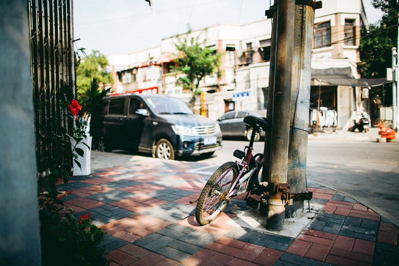 sunny corner of street with bike on sidewalk with plant and car parked on pavement, bike parked on corner of street 4k