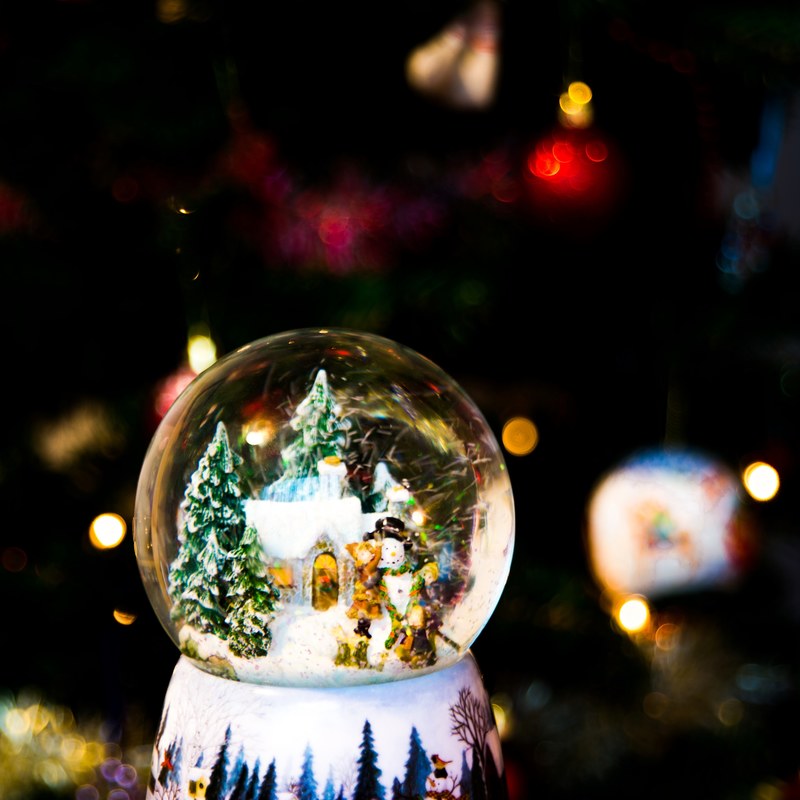 a snow globe with a winter scene displays a house surrounded by a couple evergreen trees and a snowman a decorated christmas tree is barely visible behind the snowglobe, _christmas snowglob 4k