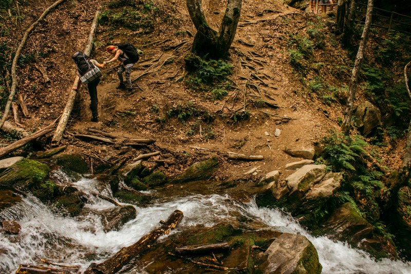 a hiker giving a hand to his friend during a descent from a steep slope by a forest creek, hiking 4k