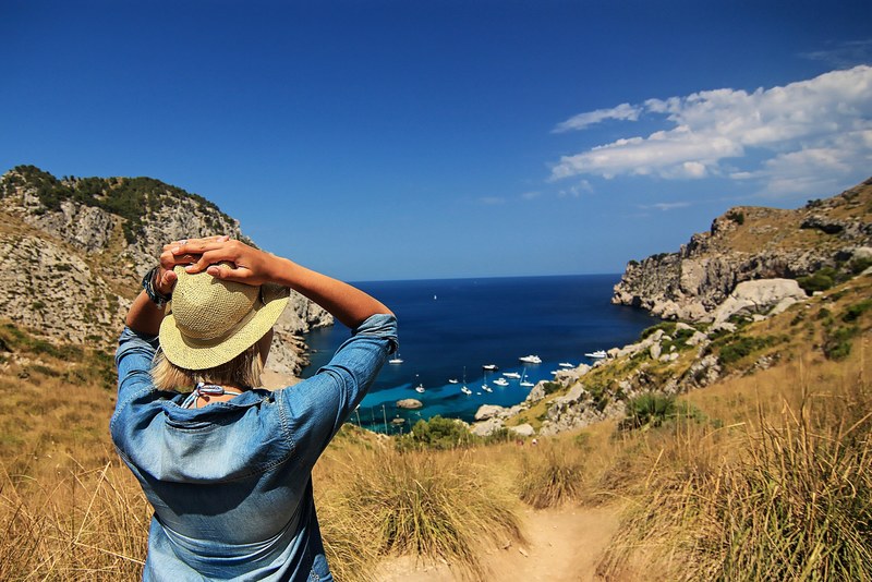 a woman in a hat and a denim jacket facing yachts in a deep blue bay encircled by cliffs, view of deep blue bay 4k