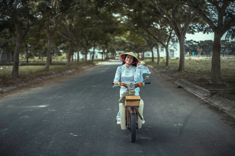 a smiling woman in a rice hat riding a vintage motorbike on a tree lined road, woman in a rice hat on motorbike 4k