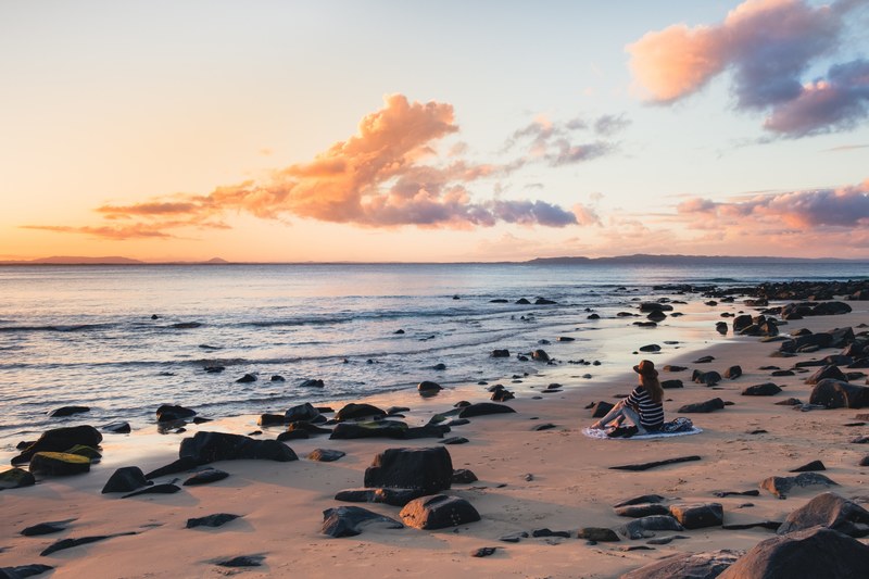 woman with hat sitting on a rocky sand beach looking at the sunset in noosa heads, noosa heads views 4k