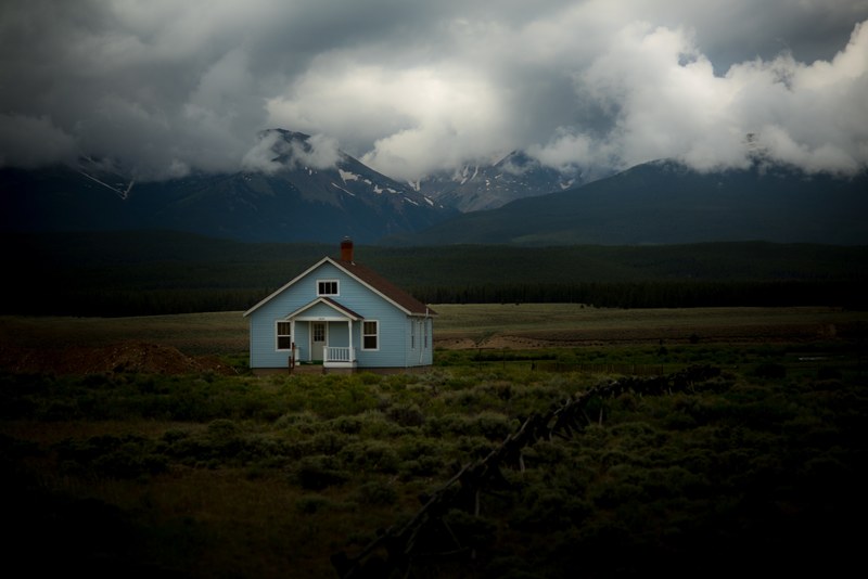 small blue house in the middle of a field of grass with mountains covered in clouds behind it, blue house in field 4k