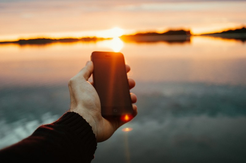 a persons hand holding a smartphone over a body of water at sunset, smartphone at sunset 4k