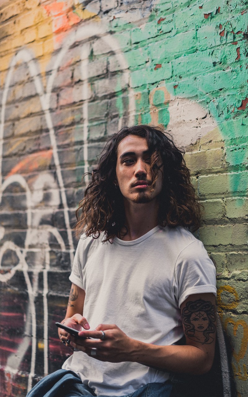a man with long curly hair leans against a graffitied wall in montreal using an iphone, whos there 4k