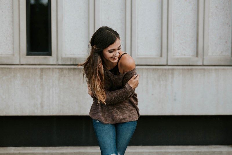 a woman in an off the shoulder sweater leans forward while laughing in front of a stone wall, woman in off shoulder sweater 4k