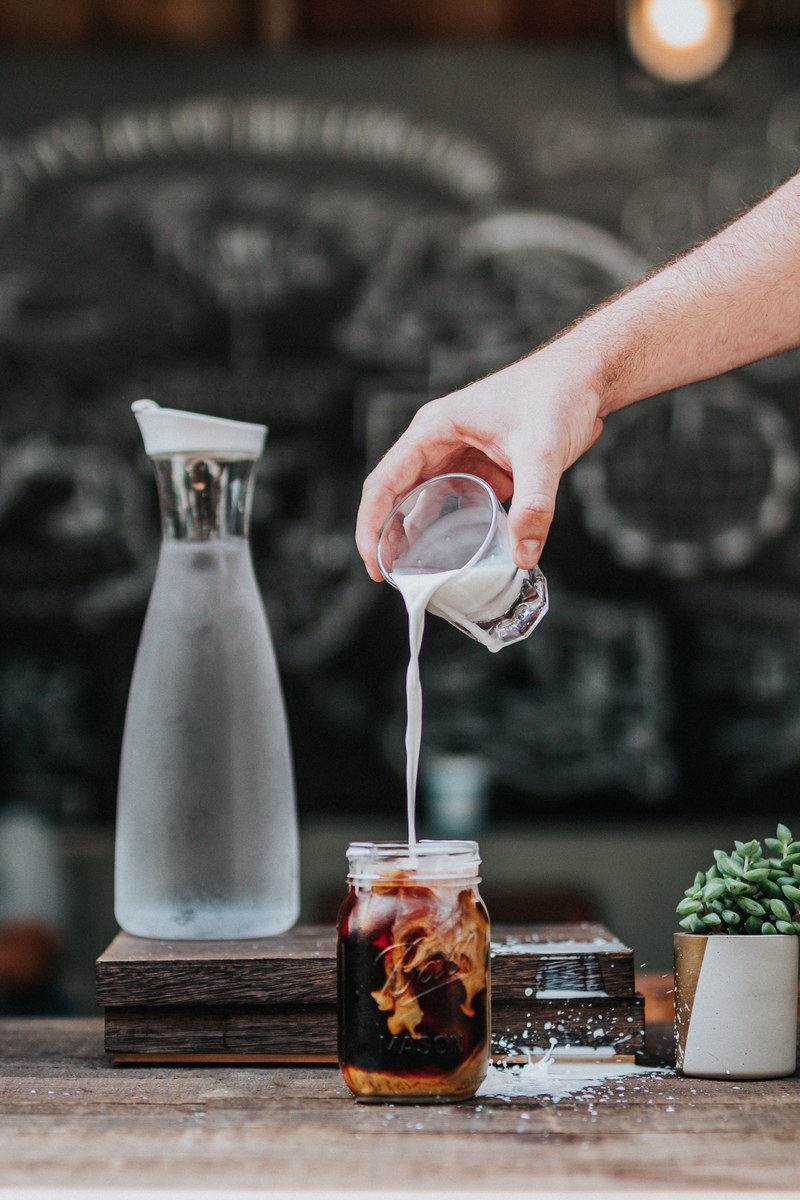 a person pouring milk into a cup of cold brew coffee in a mason jar in a cafe on a table with a water jug and succulent plant on it, cold brew coffee 4k