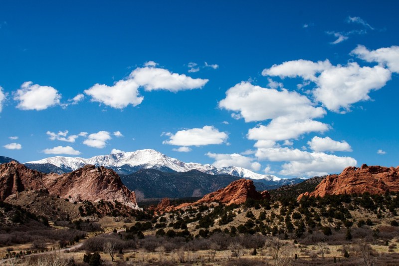 hills covered with shrubs below a red rock formation with a sunlit mountain range at the back, snowy mountains over red rocks 4k