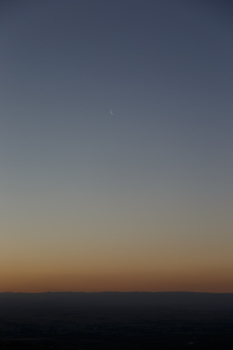 the moon like a sliver hangs on a pale blue sky at dusk over a spare landscape, moon over a spare landscape 4k