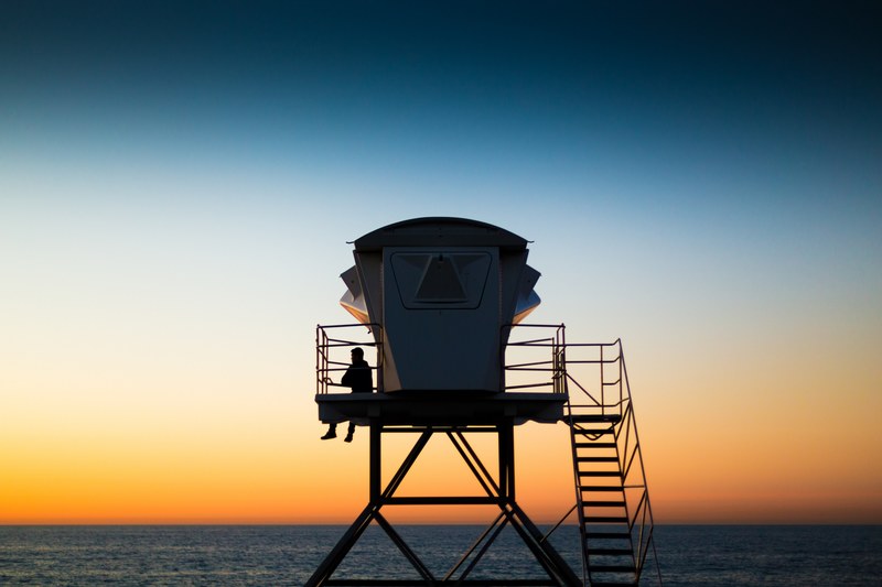 silhouette of a man sitting alone against railing of a lifeguard tower ocean and dusky sky in the background, perfect seat for a perfect sunset 4k