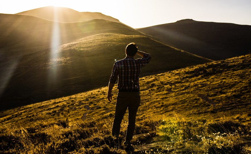a man holds his head while standing on a grassy rolling hill at sunset with lens flare effect, man hill grass and sunset 4k