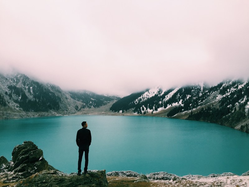 man on a rocky ledge looking at misty mountains and a lake in winter at almaty, man looking at lake almaty 4k