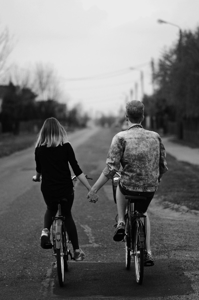 a guy and girl holding hands while cycling together on separate bikes on an empty road, lovers bike ride 4k