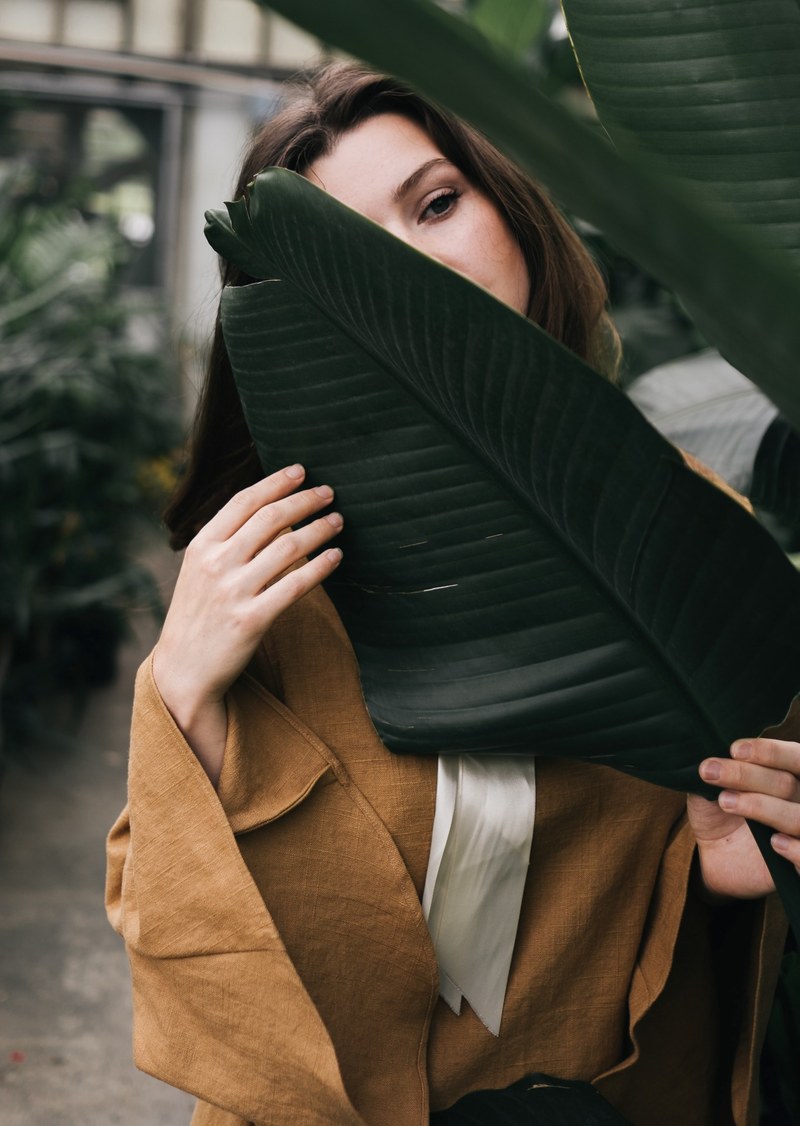 a brunette woman holding a large dark green leaf in front of her chest, brunette woman behind a leaf 4k