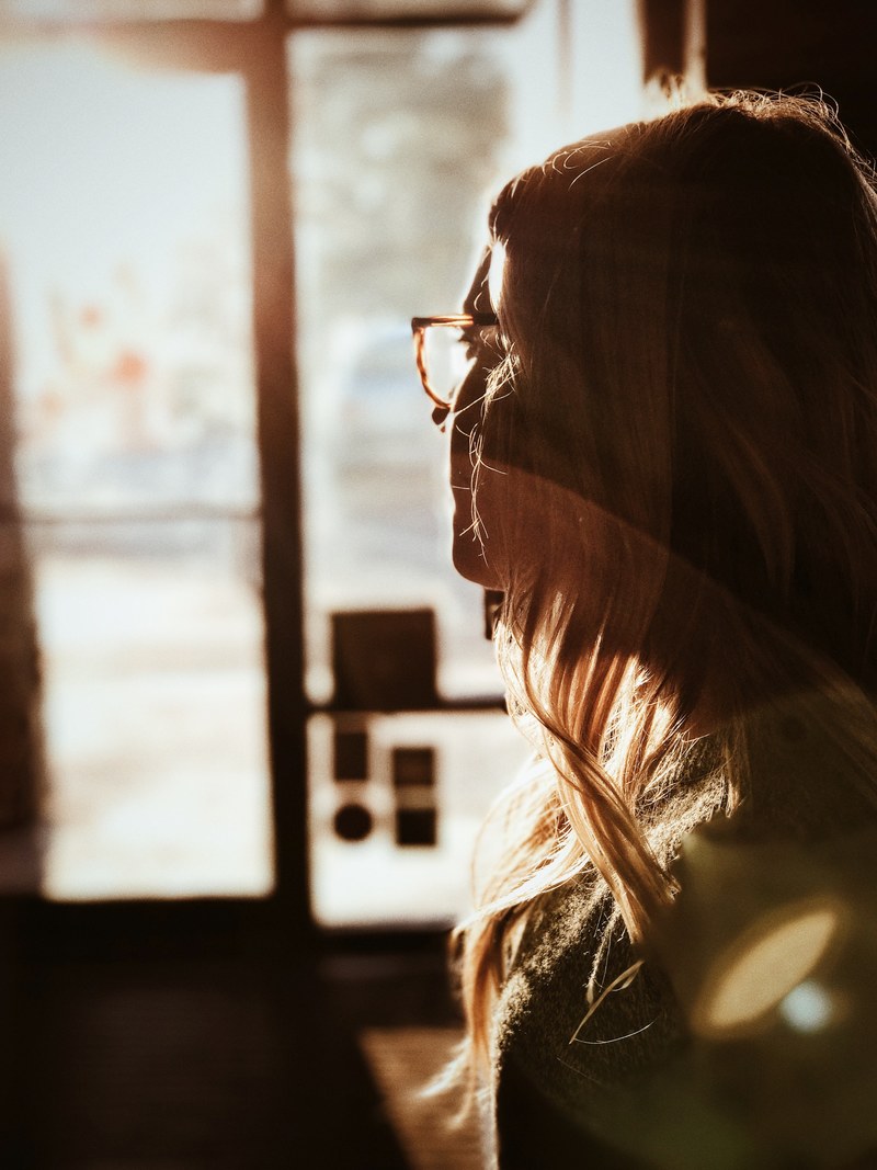 a blonde woman in glasses longingly looking at the window, woman waiting near a window 4k