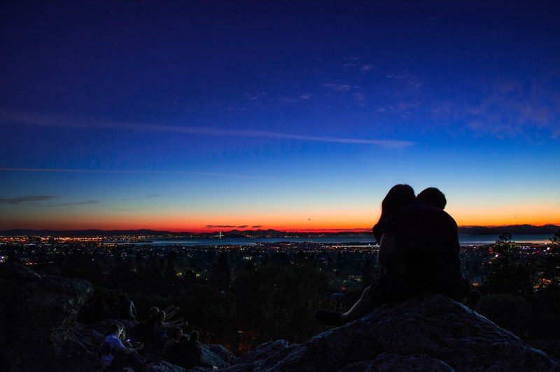 couple in silhouette kisses atop a rocky hill a city and sunset pictured in the background, kiss overlooking the sunset 4k