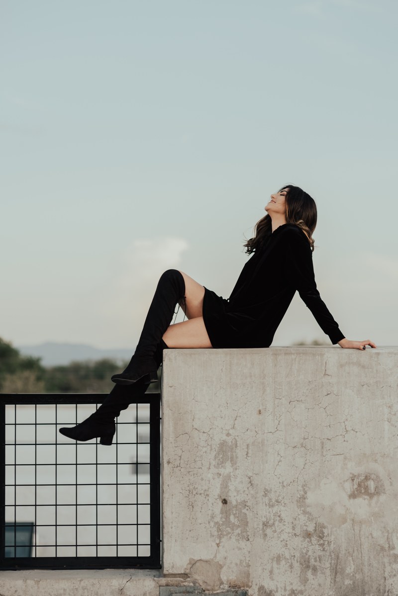 an attractive woman in a black dress with black knee high socks and high heel footwear sits posed on a cement wall in salt lake city, smile in air 4k