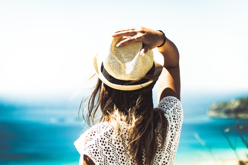 a womans back as she looks toward the ocean with one hand on her straw hat, woman looking toward ocean 4k