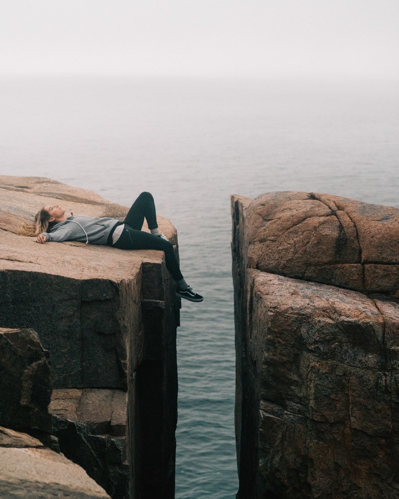 a woman laying down on her back with her feet dangling between two massive rocks over the sea, on the edge of the world 4k