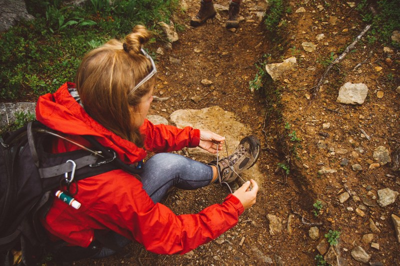 a woman in a backpack and jacket sits on dirt and rocks tying her hiking boots, woman tying hiking boots 4k
