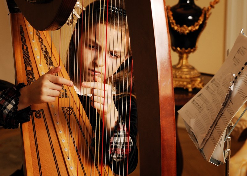 a young girl scowling as she plucks the strings of a grand wooden harp beside a sheet of music on a stand and an expensive looking vase, a young girl and her harp 4k