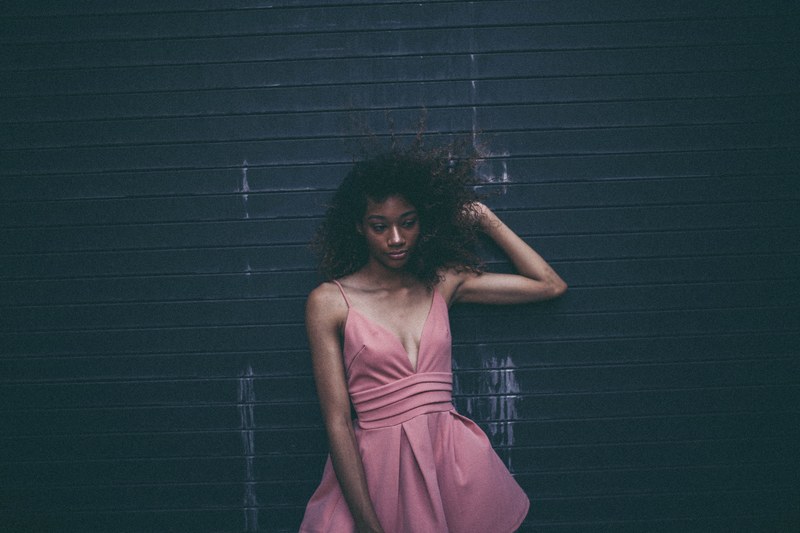 woman in a pink cocktail dress looks unsure posed against a garage door, smug face 4k