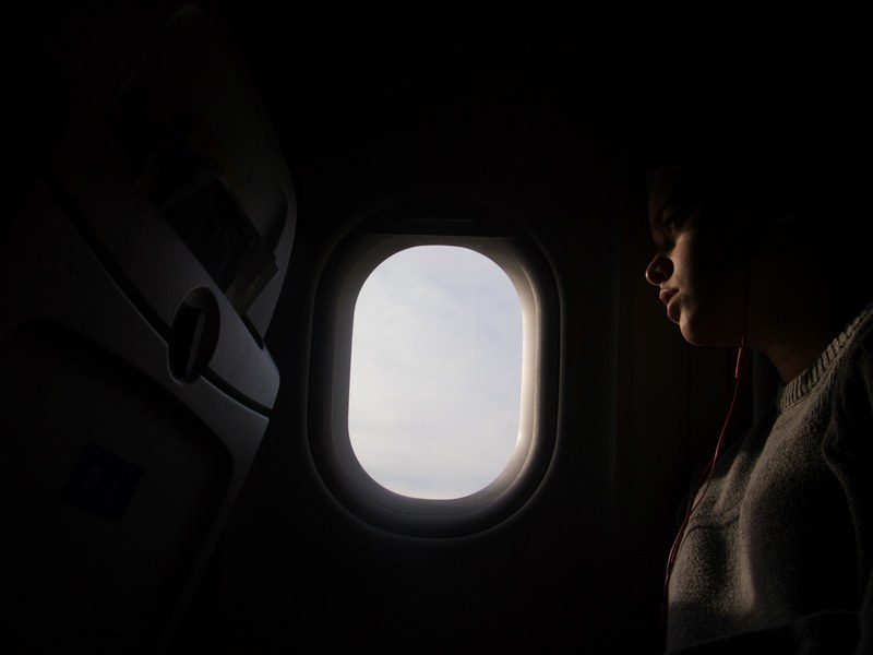 a dim shot of a person sitting next to an oval airplane window, dark plane in bright skies 4k