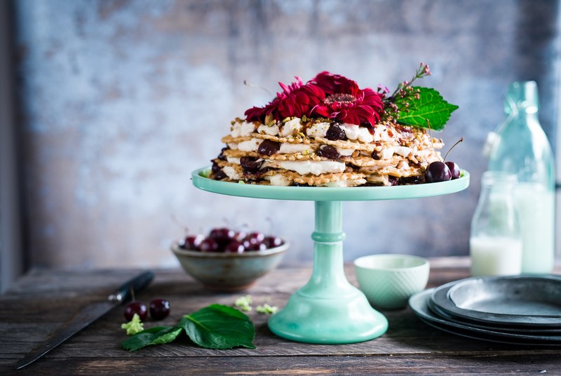 a cake stand with a cream cake covered in red flowers and cherries on a wooden table, the ugliest cake in the world 4k