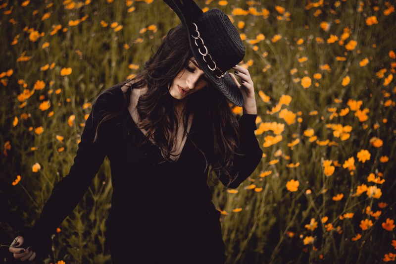 a dark haired woman in a black hat and a black dress in a field of yellow flowers, woman in black in a flower field 4k