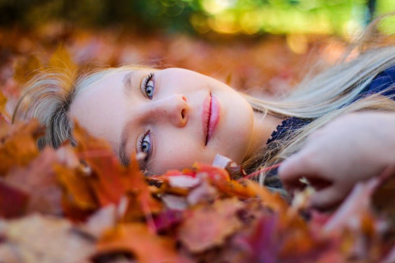 a smiling blonde woman lying in fallen leaves at the arnold arboretum of harvard university, woman lying in autumn leaves 4k