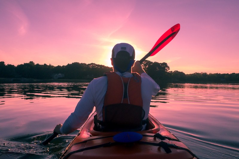 a man in a lifejacket rowing a kayak on a river at sunset, rowing into the sunset 4k