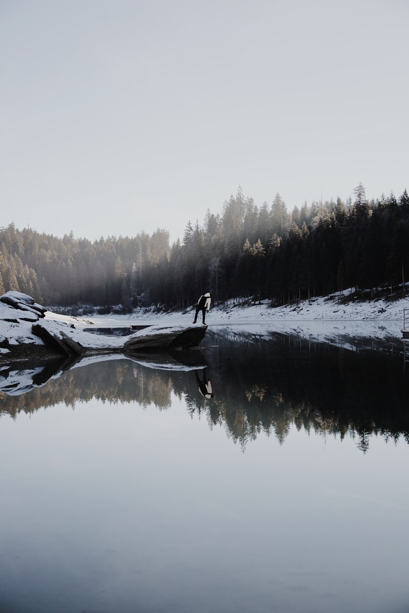 a person standing on a snow covered rock on the shore of a lake, wintry lakeside 4k