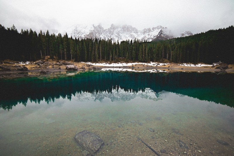 a forest of pine trees in front of a misty snowy mountain range is reflected in italys lake carezza, lake carezza forest reflection 4k